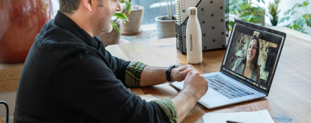 A man sits at a wooden table, using a laptop for a video call with a woman. A notebook, pen, and reusable water bottle are nearby. Plants and sunlight are visible in the background.