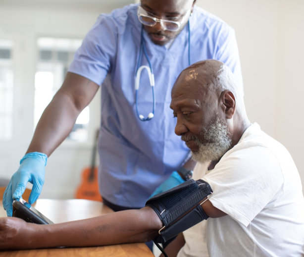 A healthcare professional in scrubs and gloves checks an elderly mans blood pressure as the man sits at a table, looking at the monitor.