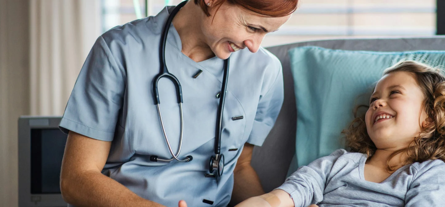 A nurse with a stethoscope smiles at a young girl lying in a hospital bed. The girl is smiling back, creating a warm and caring atmosphere.